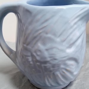 May include: A light blue ceramic pitcher with a textured, wavy design. The pitcher has a handle and a wide mouth.