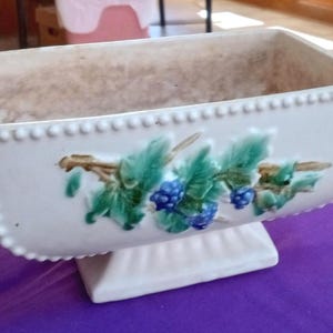May include: Rectangular white ceramic planter with a raised base. The planter is decorated with a green and blue berry and leaf design. The rim is detailed with small, raised dots. A decorative piece for plants.