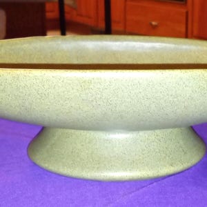 May include: A speckled, light green ceramic centerpiece bowl. The oval-shaped bowl sits on a rounded pedestal base. The bowl has a wide, shallow opening and a textured surface. The bowl is displayed on a purple surface.