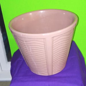 May include: A light pink ceramic planter with a wide opening and a textured, wavy design. The planter has a vertical, rectangular accent on the front. The planter is sitting on a purple surface.