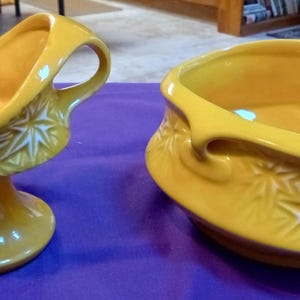 May include: Two yellow ceramic serving dishes. One is a pedestal bowl with a handle, and the other is a low bowl with a handle. Both have a starburst pattern.
