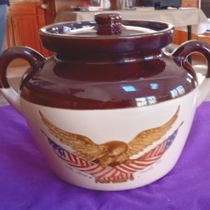 Puede incluir: Una olla de cerámica marrón y blanca con tapa y dos asas. La olla presenta un diseño de águila con banderas americanas en la parte delantera. La olla está sobre una superficie púrpura.