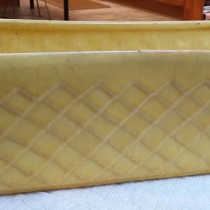 May include: A rectangular, yellow ceramic planter with a diamond pattern on the sides. The planter has a slightly flared shape and a smooth, unadorned rim. It is designed for holding plants and flowers.