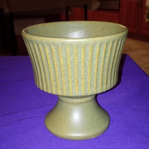 May include: A green ceramic planter with vertical ribbed detailing and a pedestal base. The planter has a speckled finish and a wide, open top. The color is a muted green with yellow accents.