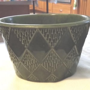 May include: A dark green ceramic planter with a diamond pattern. The planter has a wide, oval shape and a textured surface. The diamond shapes are outlined and filled with a vertical, textured design. The planter is suitable for indoor or outdoor use.