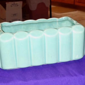 May include: A rectangular, light turquoise ceramic planter with a scalloped rim and vertical, rounded ridges. The planter is on a purple surface.