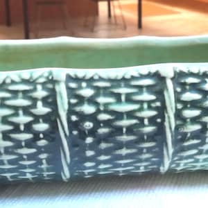 May include: A rectangular, ceramic planter with a woven basket design in shades of teal and green. The planter has a curved rim and sits on four small feet. The overall dimensions are approximately 10 inches long, 4 inches wide, and 4 inches tall.