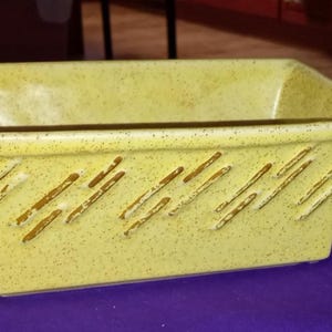 May include: A rectangular, light yellow ceramic planter with a speckled finish. The planter features diagonal brown lines on the sides. The interior is empty. A decorative piece for plants or flowers.
