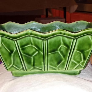 以下が含まれることがあります： ギザギザの縁が付いた、長方形の緑色のセラミックプランター。プランターの側面には幾何学的な葉の模様があります。プランターは白い布の上に置かれています。