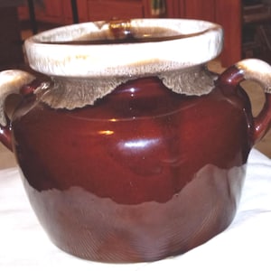May include: A vintage brown ceramic crock with a glossy finish. It features two curved handles and a wide, off-white rim. The rim has a textured, slightly rough appearance. The crock is likely used for food storage or decoration.