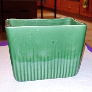 May include: A square, green ceramic planter with vertical ribbed detailing. The planter has a slightly flared rim and a glossy finish. The color is a muted, medium green, suitable for indoor or outdoor use. The planter is approximately 6 inches wide and 6 inches tall.