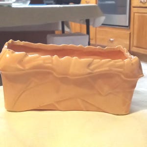 May include: A peach-colored ceramic planter shaped like a paper bag. The rectangular planter has a textured surface with folds and creases, and a smooth interior. The open top reveals the inside of the planter.
