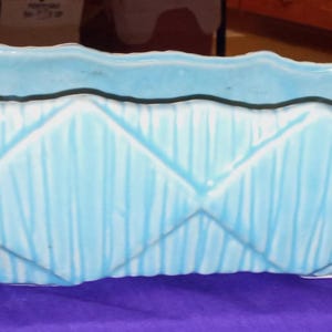 以下が含まれることがあります： 長方形の形とテクスチャ加工された幾何学模様が特徴の、水色のセラミックプランター。 プランターは波状の上端を持ち、紫色の表面に置かれています。