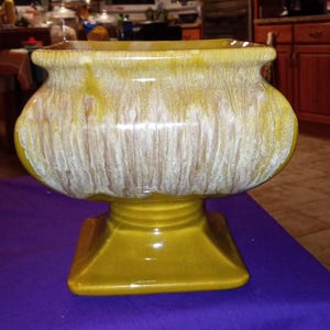 May include: A yellow ceramic planter with a square base and a bulbous body. The planter has a textured, dripped white and yellow glaze. The overall shape is reminiscent of a pedestal bowl.