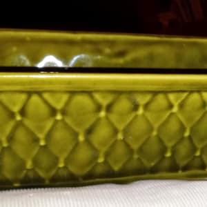 May include: A rectangular, olive green ceramic planter with a diamond pattern on the sides. The planter has a raised rim and sits on small feet. The interior is dark, suggesting it is empty. The overall design is vintage.
