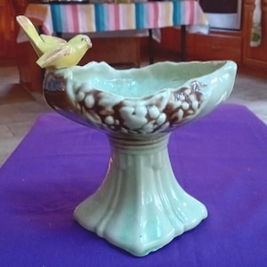 May include: A ceramic pedestal bowl with a light green base and a brown and white rim. A yellow bird figurine is perched on the edge. The bowl has a scalloped edge and a textured surface. The bowl is likely for decorative purposes.