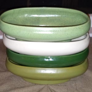 May include: A stack of four ceramic bowls in shades of green and white. The top bowl is a light green, followed by white, dark green, and a muted green at the bottom. The bowls are oval-shaped.