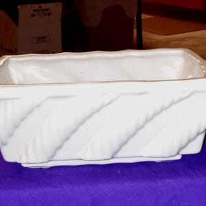 Op de afbeelding: Een rechthoekige witte keramische plantenbak met een gestructureerd, diagonaal touwachtig ontwerp aan de zijkanten. De plantenbak heeft een eenvoudige, open bovenkant en staat op een platte basis. Ideaal voor binnen of buiten.