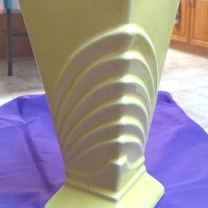 May include: A pale yellow ceramic vase with a geometric design. The vase has a square base and a flared, angular body. A series of curved, fan-like ridges adorn one side. The vase is set against a purple cloth.