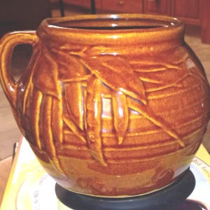 May include: A brown ceramic mug with a handle and a textured bamboo leaf design. The mug has a wide opening and a rounded body. The exterior is glazed with a glossy finish. The mug is likely used for beverages.