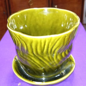 May include: A green ceramic planter with a matching saucer. The planter has a wavy, textured design and a glossy finish. The saucer has a small drainage hole. The overall shape is round and the color is a deep, olive green.