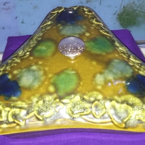 May include: A triangular-shaped decorative dish with a yellow and gold glaze. The dish features blue and green accents and a central silver-colored, circular embellishment. The edges have a textured, ornate design.