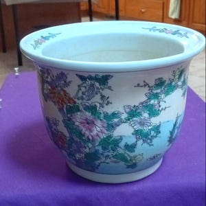 May include: A white ceramic planter featuring a wide, rounded rim and a tapered base. The exterior is decorated with a colorful, detailed illustration of blooming flowers, green foliage, and branches in shades of pink, orange, and green.
