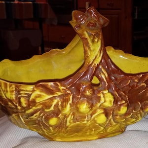May include: A ceramic basket with a handle, featuring a yellow interior and a brown exterior with a leaf and berry design. The basket has a textured surface and is likely used for decorative purposes or storage.