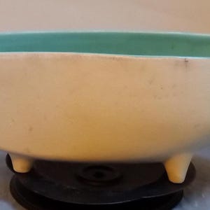 May include: A white ceramic bowl with a light green interior and three legs. The bowl is oval shaped and has a smooth, glossy finish.