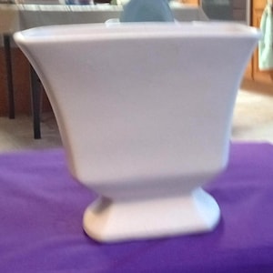 May include: A white, vase-shaped container with a flared top and a pedestal base. The container is sitting on a purple surface. The vase has a smooth, matte finish and a simple, elegant design.