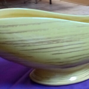 May include: A large, yellow ceramic bowl with a pedestal base. The bowl has a boat-like shape with a wide opening and a textured surface with brown horizontal lines. The bowl is likely for decorative purposes.