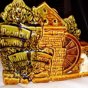 以下が含まれることがあります： 水車小屋のシーンを描いたセラミック製の置物。茶色と緑の色合いで、水車、小さな家、そして「Down by the old Mill Stream」の文字が特徴です。