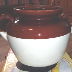 Puede incluir: Un tarro de cerámica con dos asas, de diseño clásico. La parte superior es marrón oscuro, la sección media es blanca y el borde y las asas son marrones. Este recipiente de estilo vintage es adecuado para la cocina o la decoración del hogar.