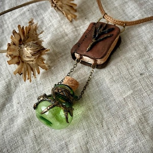 May include: A necklace featuring a brown rectangular pendant resembling a small book with a dark branch-like detail, connected by thin metal chains to a small, round, translucent green glass bottle with a cork stopper and dark metal accents.