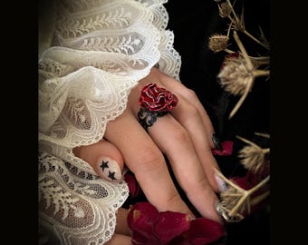Peut inclure: Une bague en forme de rose rouge avec des détails en filigrane noir. La bague est portée sur un doigt avec un art d'ongles en forme d'étoile noire.