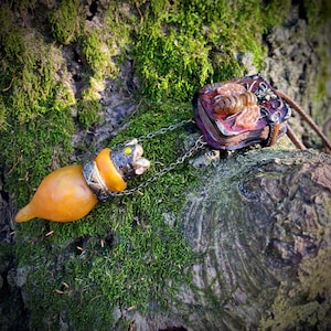 May include: A necklace with two charms: a small, orange, ceramic, animal-shaped charm and a small, red, book-shaped charm with a bee design. The charms are attached to a silver chain.