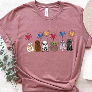 May include: A dusty rose t-shirt featuring a graphic of Star Wars characters holding heart-shaped balloons. The characters include BB-8, Chewbacca, a Stormtrooper, Yoda, and Darth Vader. A beige hat and white mug are also in the image.