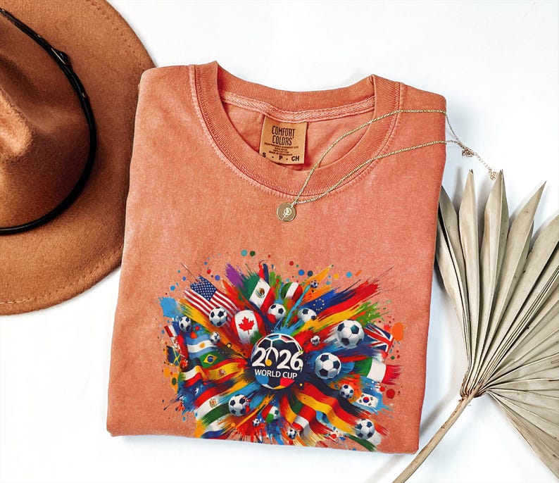 May include: A coral-colored Comfort Colors t-shirt with a crew neck and short sleeves. The shirt features a colorful graphic of flags and soccer balls, with the text "2026 World Cup". A gold necklace and a brown hat are also in the image.