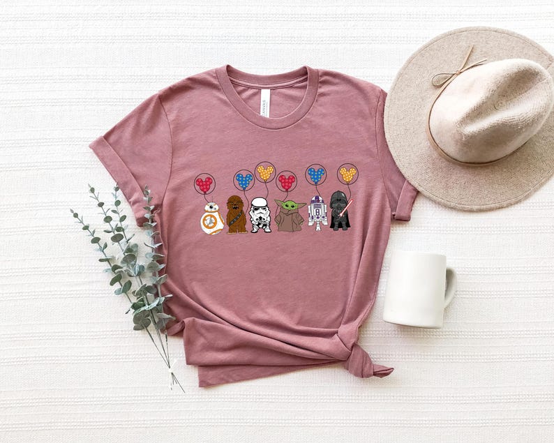 May include: A dusty rose t-shirt featuring a graphic of Star Wars characters holding heart-shaped balloons. The characters include BB-8, Chewbacca, a Stormtrooper, Yoda, and Darth Vader. A beige hat and white mug are also in the image.