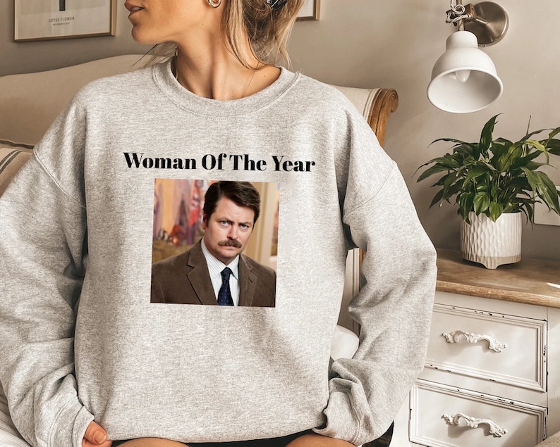 May include: Gray sweatshirt with the text "Woman Of The Year" and a picture of Ron Swanson from the TV show Parks and Recreation.