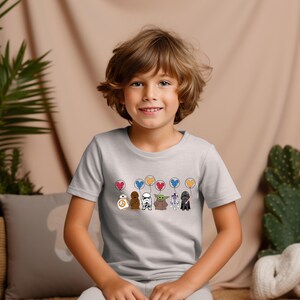 May include: A light gray t-shirt featuring cartoon Star Wars characters holding heart-shaped balloons. Characters include BB-8, Chewbacca, a Stormtrooper, Grogu, Darth Vader, and R2-D2. The balloons are red, blue, and yellow.