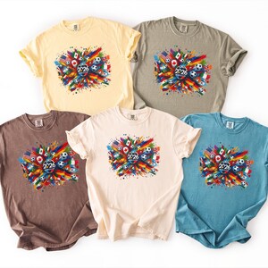 May include: Five short-sleeved t-shirts in various colors: yellow, grey, brown, cream, and blue. Each shirt features a colorful graphic with flags, soccer balls, and the number "2026". The design is centered on the front of each shirt.