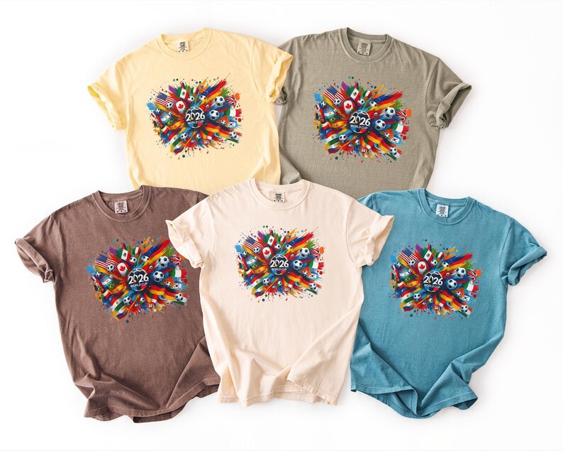 May include: Five short-sleeved t-shirts in various colors: yellow, grey, brown, cream, and blue. Each shirt features a colorful graphic with flags, soccer balls, and the number "2026". The design is centered on the front of each shirt.