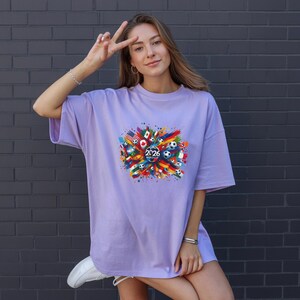 May include: A light purple t-shirt with a colorful graphic design featuring flags, soccer balls, and the year "2026". The design is centered on the front of the shirt. The shirt has short sleeves and a relaxed fit.
