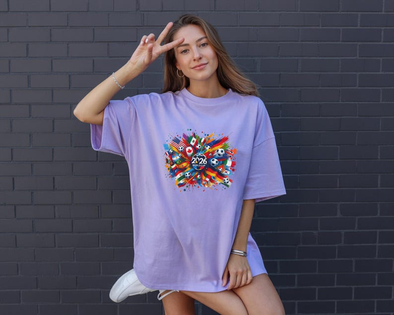May include: A light purple t-shirt with a colorful graphic design featuring flags, soccer balls, and the year "2026". The design is centered on the front of the shirt. The shirt has short sleeves and a relaxed fit.