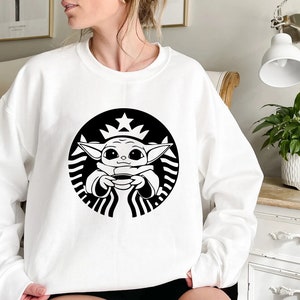 May include: White sweatshirt with a black and white graphic of a cartoon character holding a cup of coffee. The character is inside a circle with a star on top and a wavy line around the edge. The graphic is a parody of the Starbucks logo.