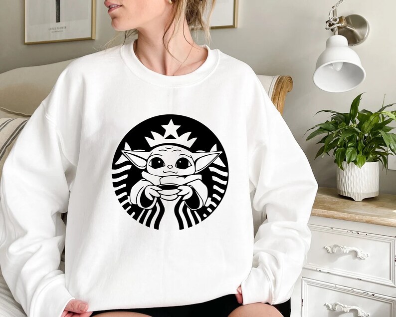 May include: White sweatshirt with a black and white graphic of a cartoon character holding a cup of coffee. The character is inside a circle with a star on top and a wavy line around the edge. The graphic is a parody of the Starbucks logo.