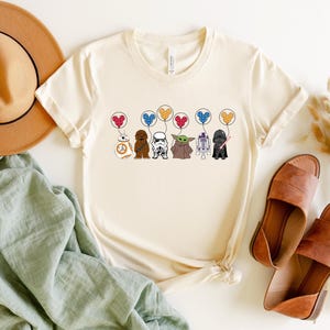 May include: Cream-colored t-shirt featuring cartoon Star Wars characters holding heart-shaped balloons. Characters include BB-8, Chewbacca, a Stormtrooper, Yoda, R2-D2, and Darth Vader. The shirt is paired with brown sandals, a tan hat, and a green cloth.