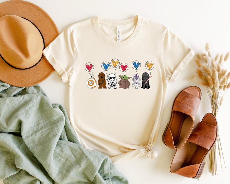 May include: Cream-colored t-shirt featuring cartoon Star Wars characters holding heart-shaped balloons. Characters include BB-8, Chewbacca, a Stormtrooper, Yoda, R2-D2, and Darth Vader. The shirt is paired with brown sandals, a tan hat, and a green cloth.