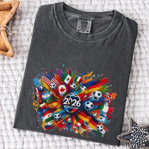 May include: Dark gray t-shirt featuring a colorful graphic design for the 2026 World Cup. The design includes soccer balls and flags from various countries, with the text "2026 WORLD CUP" in the center.